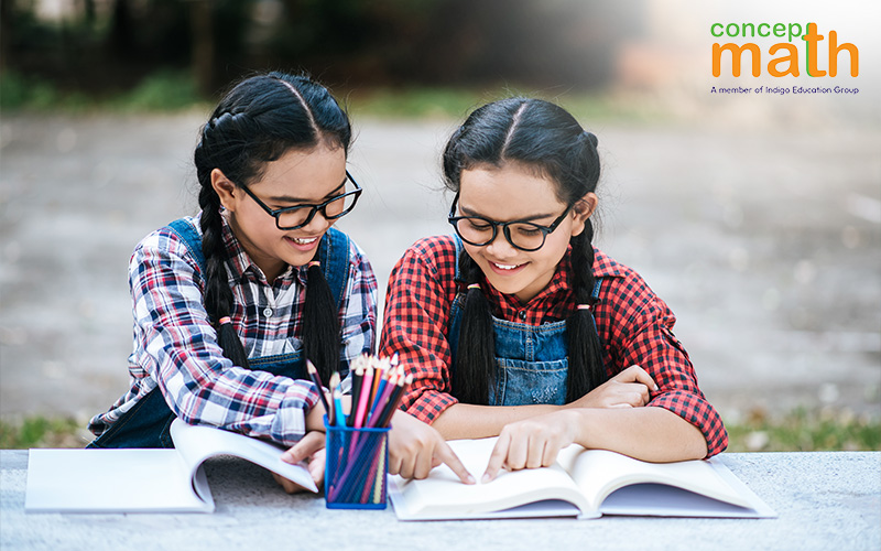 Two smiling girls with braided hair and glasses read a book together outdoors.